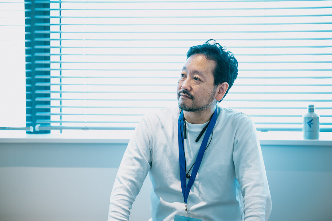 奥にブラインドのついた窓のある壁を背にして座っている